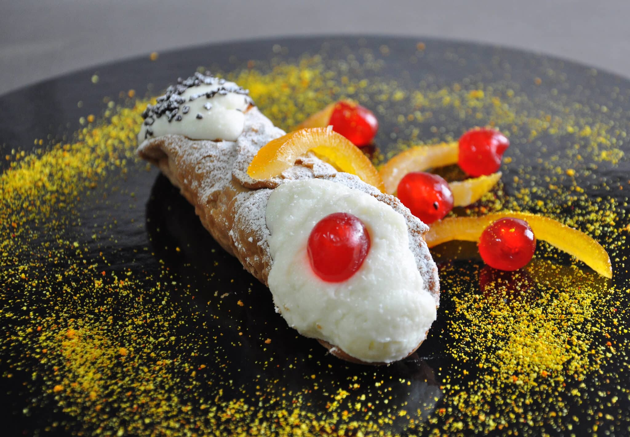 Cannolo siciliano servito su un piatto con ripieno di ricotta, scorze d'arancia candite su piatto nero con ciliegie candite. Arte dolciaria siciliana, Italia.